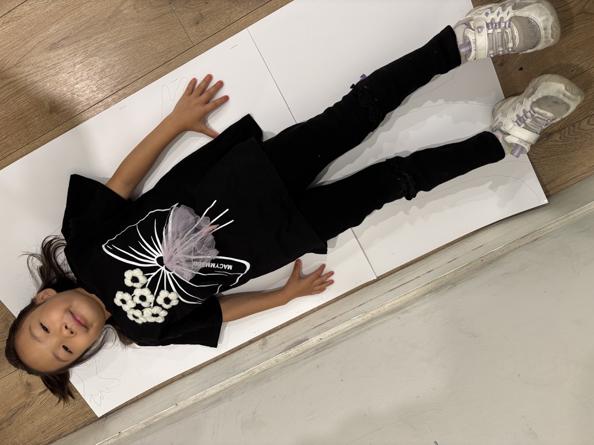 A student lying still on a long sheet of white paper on the floor while her body outline is traced around her