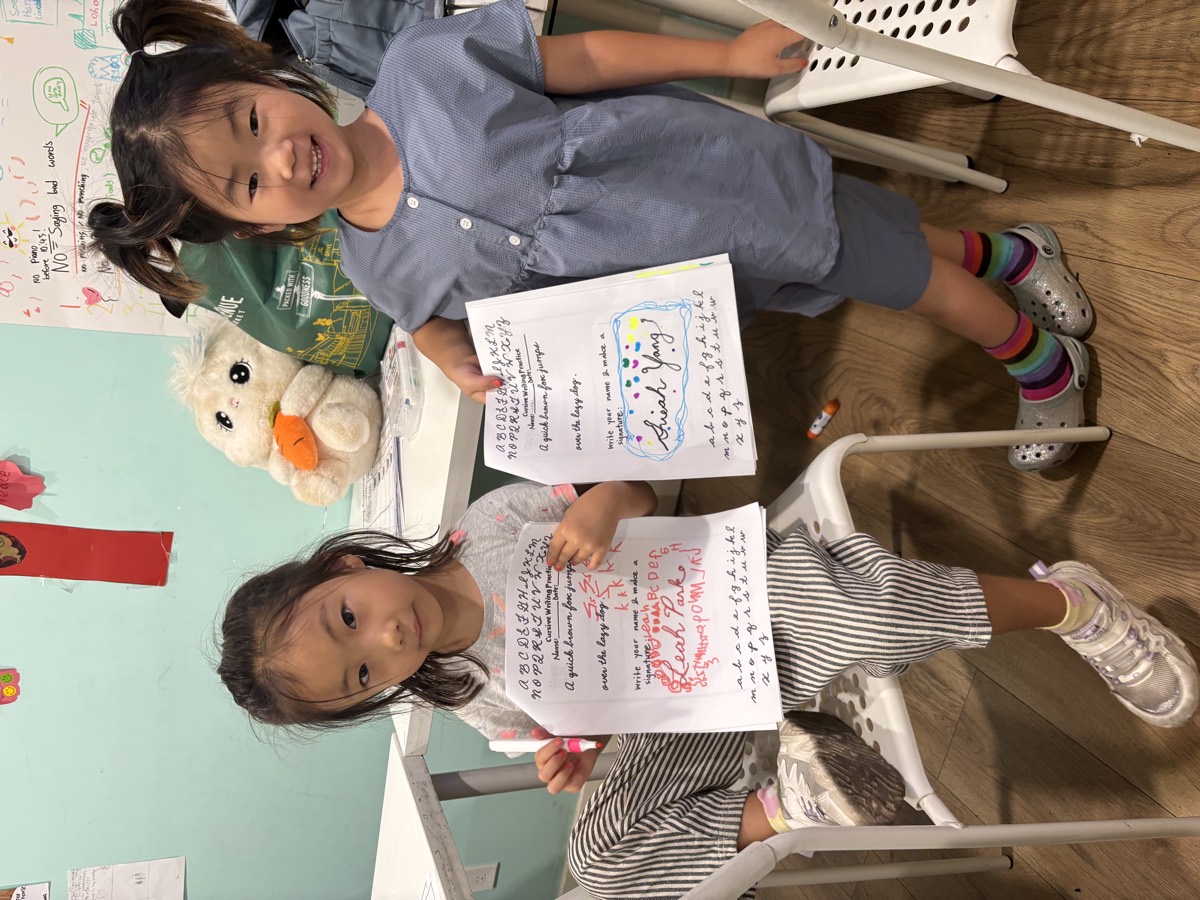 Two young students holding up their completed cursive writing practice sheets, each with their own signature decorated in colour
