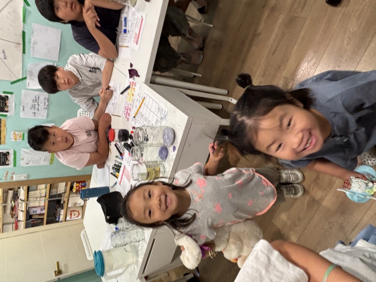 A young student grinning at the camera with the jars of coloured liquid lined up on the table behind her