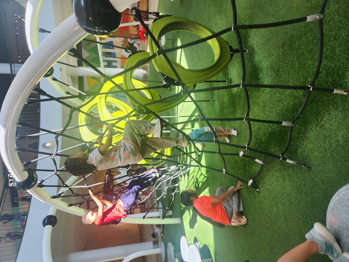 Children climbing a black rope net over green turf at the indoor playground