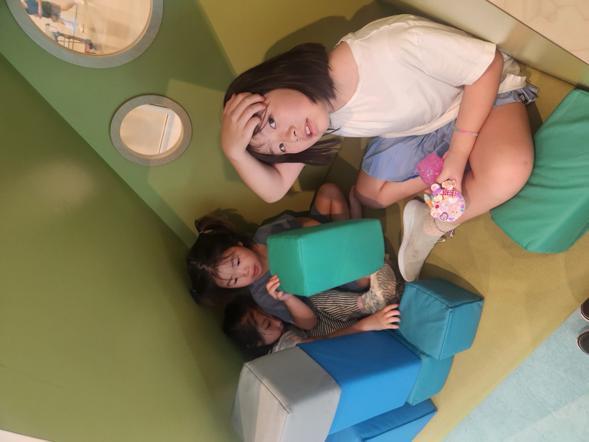 Three children sitting together inside a small green and blue play pod with foam blocks