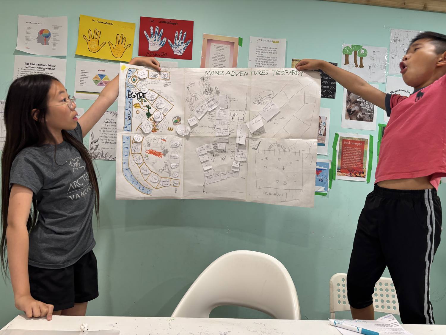 Two students presenting their hand-drawn 'Moses Adventures Jeopardy' board game