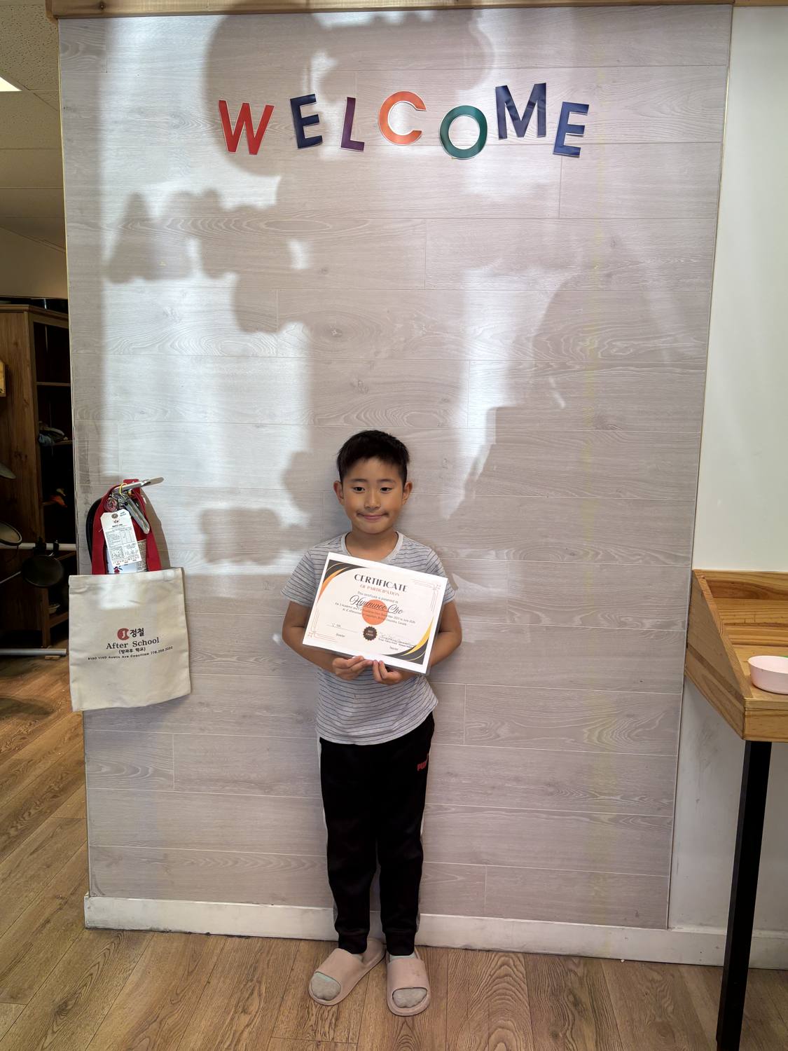 Hyunwoo holding his certificate of completion under the WELCOME wall